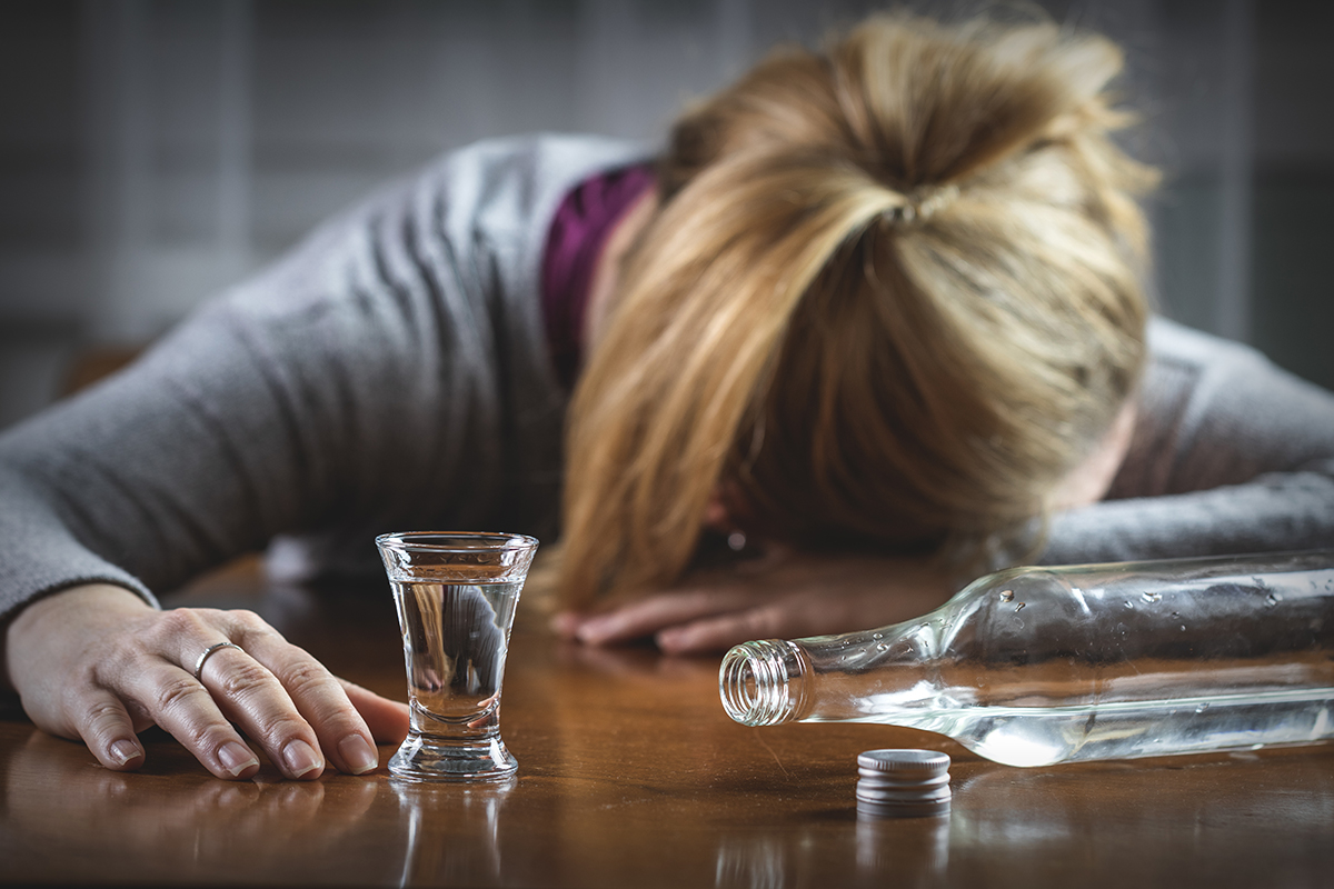 a woman puts her head down in front of a bottle of clear liquid as she considers the link between alcohol and depression