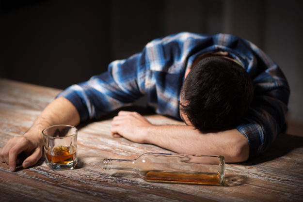 a man puts his head on a table next to a bottle of alcohol as he wonders if he has an alcohol dependence