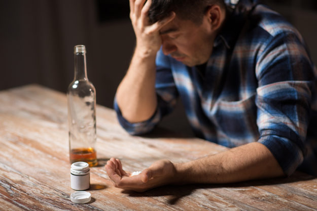 a man sits in front of a bottle of alcohol and a pill bottle and considers taking benzos and alcohol together
